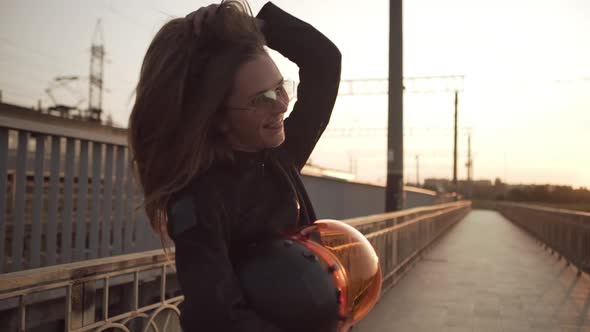 Smiling Moto Girl in Black Leather Jacket and Sunglasses Wearing Braces Posing on Footbridge Looking