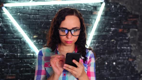 Young Beautiful Woman with Glasses Stands with Mobile Phone in Hands on Background Brick Wall 