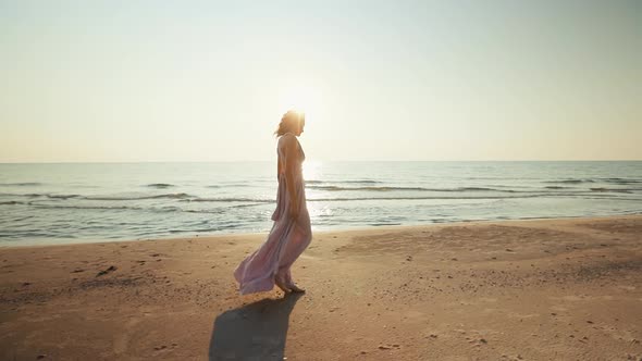 Panning Pretty Woman in Long Dress Walking on Wet Sand Against Sunset Sea Background
