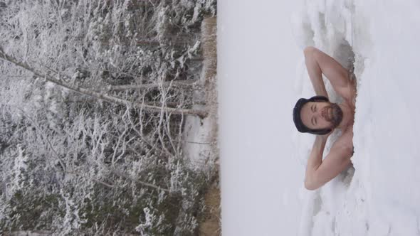 VERTICAL - A handsome, rugged hipster man relaxes in his ice hole facing camera