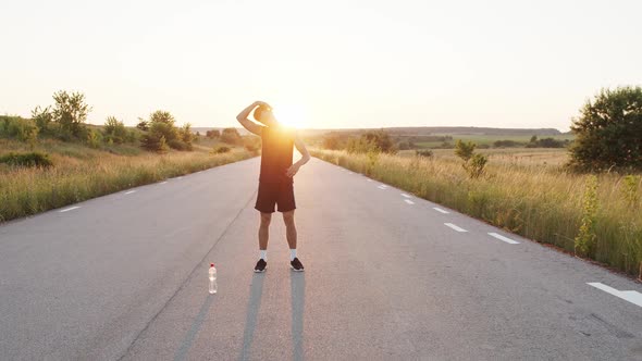 Young Athlete Warming Body Up on Country Road with Bottle of Water at Sunset