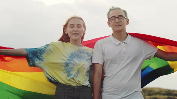 A Young Couple Holds Hands and Raises a Rainbow LGBT Flag While Looking at the Camera