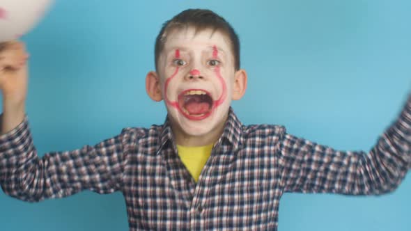 Caucasian boy 7-8 years old in scary clown makeup hides behind balloons scares looking at the camera