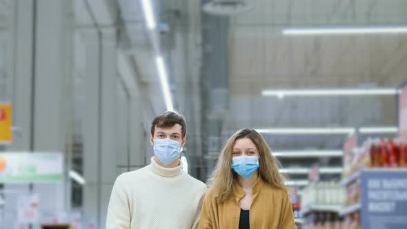 Young Married Couple in Medical Masks and Gloves Stand in Supermarket with Grocery Cart, Camera