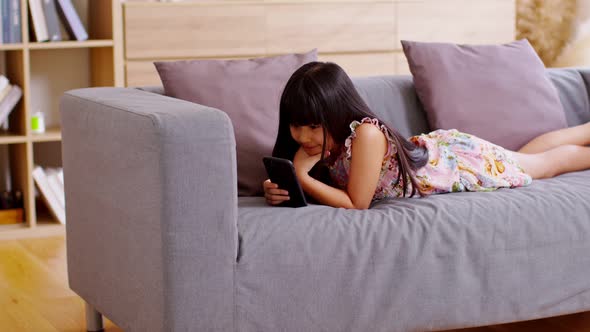 Cute little girl uses smartphone while prone at the sofa in the living room. Child surfing the inter