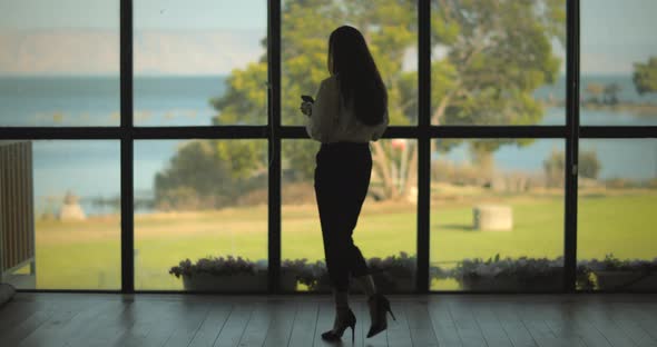 Businesswoman checking her phone near a big window with a lake view. Slow motion, long shot.