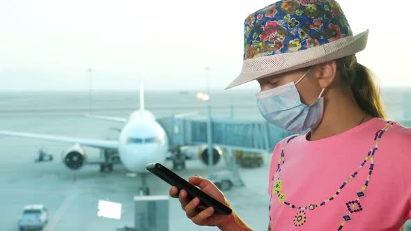 Teenage Girl, in Protective Mask, Uses Mobile, in Front of Panoramic Window with Runway 