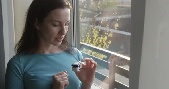 Pretty Female Is Using Pulse Oximeter Checking Her Oxygen Level Standing By The Window