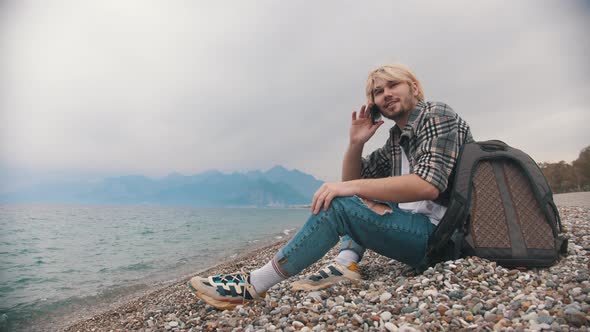 A Young Man in a Plaid Shirt Sits on the Shore and Talks with His Friend on the Phone