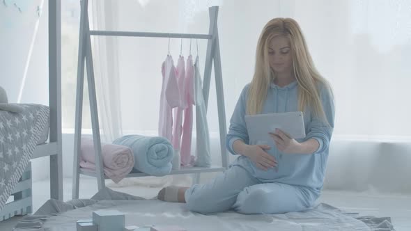 Portrait of Happy Pregnant Woman Using Tablet and Touching Kids Clothes Hanging on Hanger. Smiling