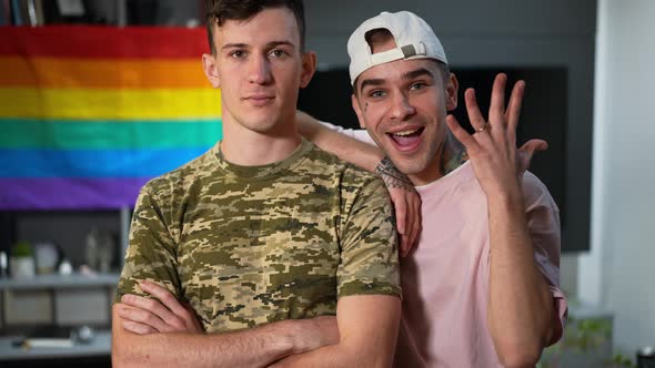 Portrait of Excited Young Gay Man Boasting Engagement Ring Standing with Military Boyfriend Indoors