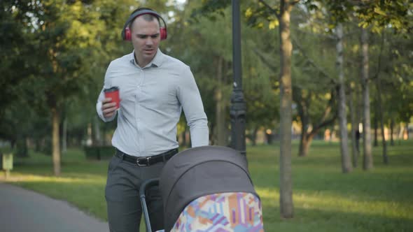 Portrait of Positive Young Man in Headphones Pushing Baby Stroller in Summer Park, Singing and