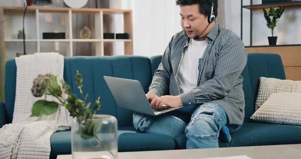 Asian Man in Headset Sitting on Comfortable Sofa in Home Office and Working on Laptop