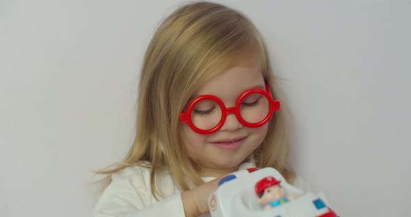 Child Girl in Red Glasses Plays at the Doctor with Toy Ambulance.