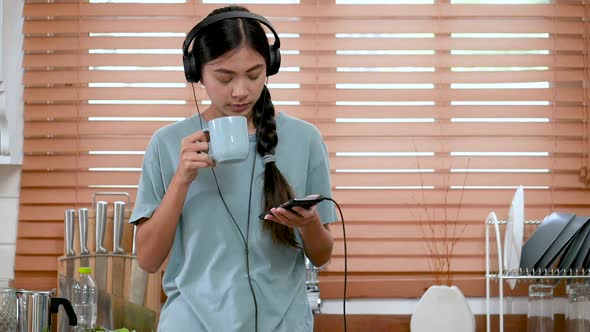 Young Asian woman listening to music on smartphone and drinking coffee in the kitchen at home.