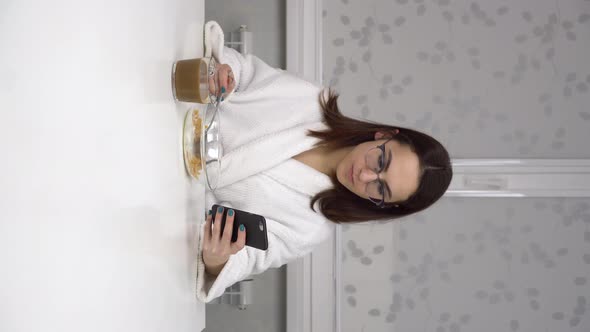 A Cheerful Young Woman in a White Coat Eats Cereals and Drinks Coffee and Uses a Smartphone