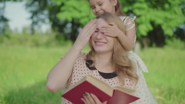 Charming Little Girl Coming To Mom Reading Book in Park and Closing Her Eyes. Portrait of Positive