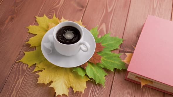 Coffe Cup on Wooden Table with Book and Colorful Autumnal Maple Leaves with Spinning Coffee Bubbles