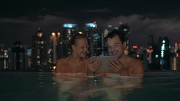 Couple With Touch Pad In Rooftop Pool At Night