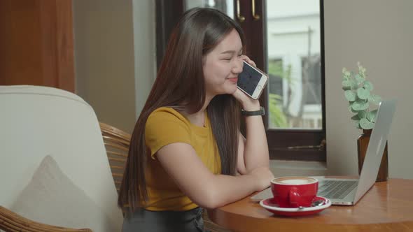 Happy Young Asian Girl Talking on Phone at a Coffee Shop with a Laptop