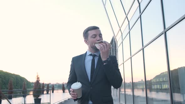 Young Handsome Businessman Holding a Cup of Coffee Eating a Burger on the Street