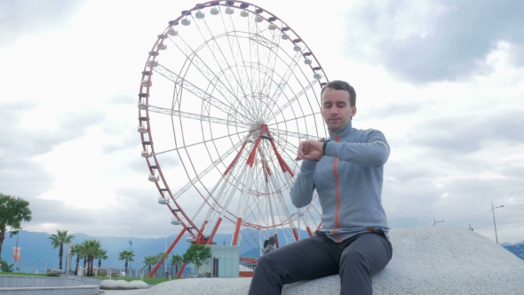 A Young Man Checks The Messages On The Smart Watch Around The Ferris Wheel. Early Morning. The Wheel