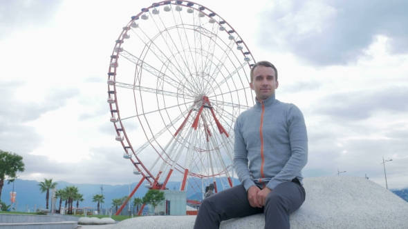 Young Handsome Man Looking Into The Camera And Smiles Around The Ferris Wheel. Early Morning. The