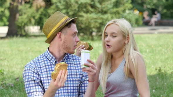 Young Couple Eating Sandwiches on a Lawn Talking