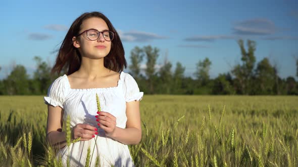 A Young Woman in a White Dress Stands in a Green Wheat Field and Holds a Spikelet in Her Hands. The