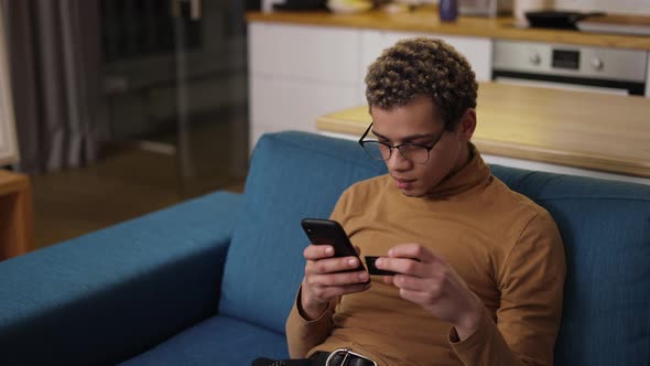 Happy Young Man Buying Online Uses a Phone and a Credit Card on Sofa Home