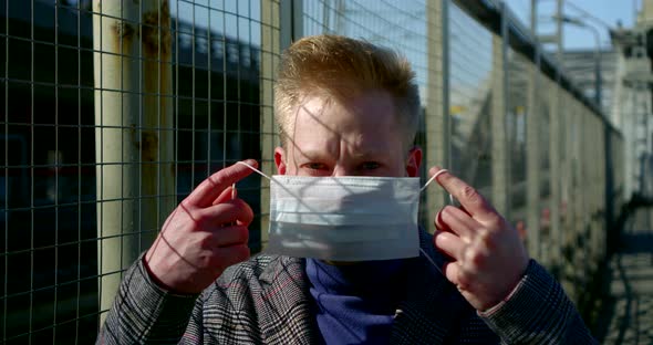 Portrait of a Man on a City Street. He Stands Near the Metal Fence Grating, Puts a Disposable Mask