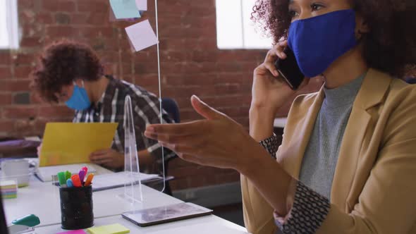 Mixed race businesswoman talking on phone sitting in front of computer wearing face mask