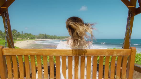 Woman in Dress Swinging with Tropical Beach View While Wind Blowing Blonde Hair