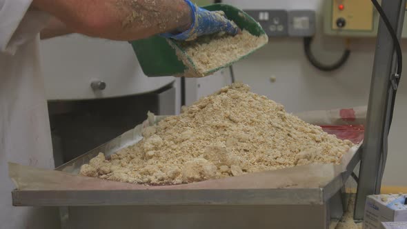 A worker add measures of crumble mix to a tray pf cake mix, res some due to it being overweight