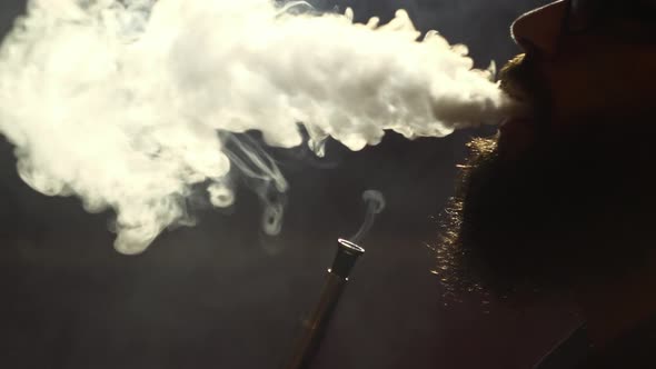 Young Bearded Man in Glasses Smokes a Hookah and Blow Out Smoke Closeup on Black Background in Slow