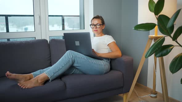 Woman with Glasses is Sitting on the Couch and Working on a Laptop