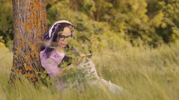 Young Woman in Glasses Headphones Reading Book Sitting Near Tree in Summertime