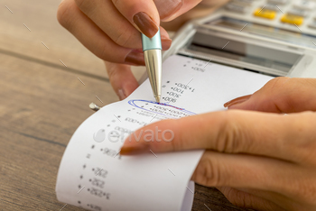  female hands working with adding machine and printout circling important figures wit a pen.