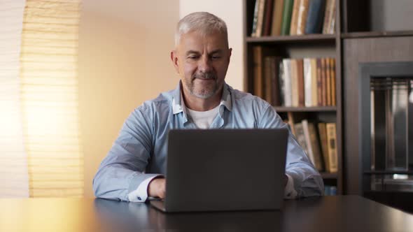 Mature Man Surfing Internet on Laptop Computer Sitting at Cabinet at Home