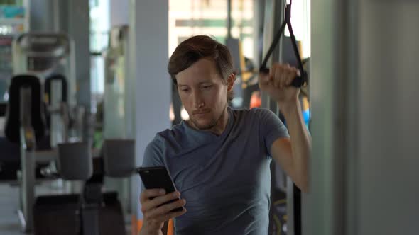 A Young Man at the Gym Doing Exercises Holding a Phone in Front of His Eyes in His Hands. All the