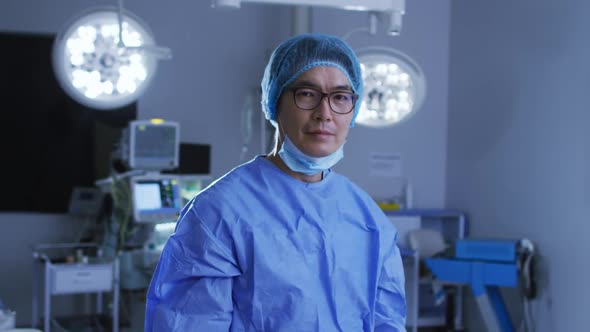 Portrait of asian male surgeon wearing lowered face mask, crossing arms in operating theatre