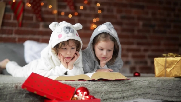 Two Little Cute Siblings Reading a Book In Bed Near Christmas Tree With Lights And Illumination