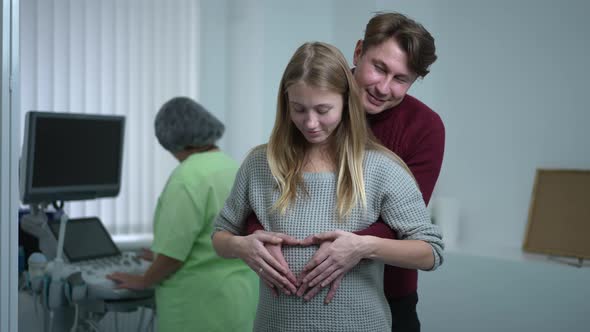 Happy Caucasian Couple Gesturing Heart Shape on Pregnant Belly Looking at Camera Smiling