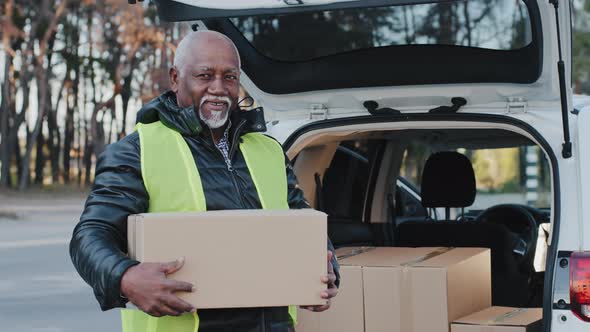 Mature African American Smiling Man Holding Cardboard Box Stands Near Open Trunk of Automobile Happy