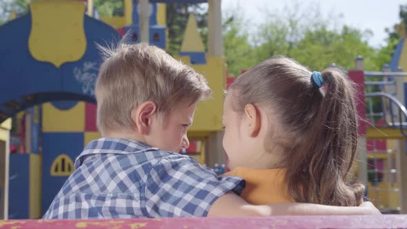 Cute Blond Boy and a Pretty Girl Sitting on the Bench in Front of the Playground Hugging. A Couple