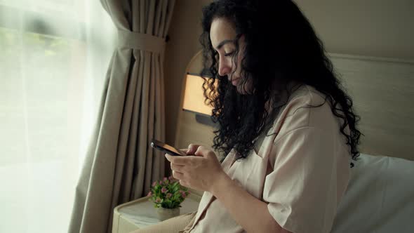 Cheerful Young Caucasian Woman Using Smartphone While Sitting on Bedroom