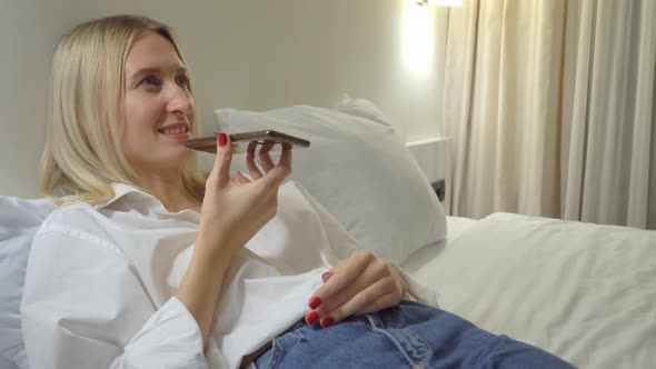 a woman at home records a voice message on her smartphone while lying on bed.