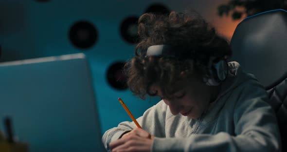 A Boy with Curly Hair Sits in a Room at a Desk with a Laptop on Wireless Headphones Taking Notes