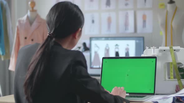 Back View Of Asian Female Designer In Business Suit Using A Laptop With Mock-Up Green Screen