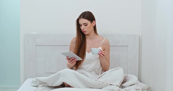 Female Freelancer Is Sitting on a Bed Drinking Coffee and Working on a Screen Tablet, Morning Time